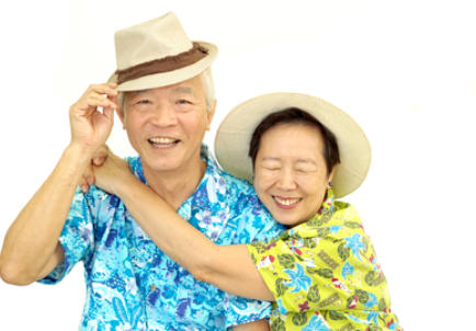 A senior couple on a white background. They are wearing Hawaiian shirts and sun hats. They're both smiling, and the woman is hugging the man with her head resting on his shoulder.