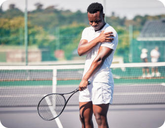 A young man stands on a tennis court holding a racquet in his left hand. His right hand is holding his left shoulder and he is grimacing in pain.