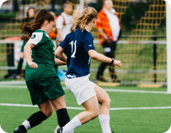 Two teenage girls wearing different soccer uniforms are on the soccer pitch both trying to kick the ball.