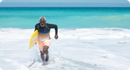 A senior man is running out of the waves onto the beach carrying a surfboard under his arm.